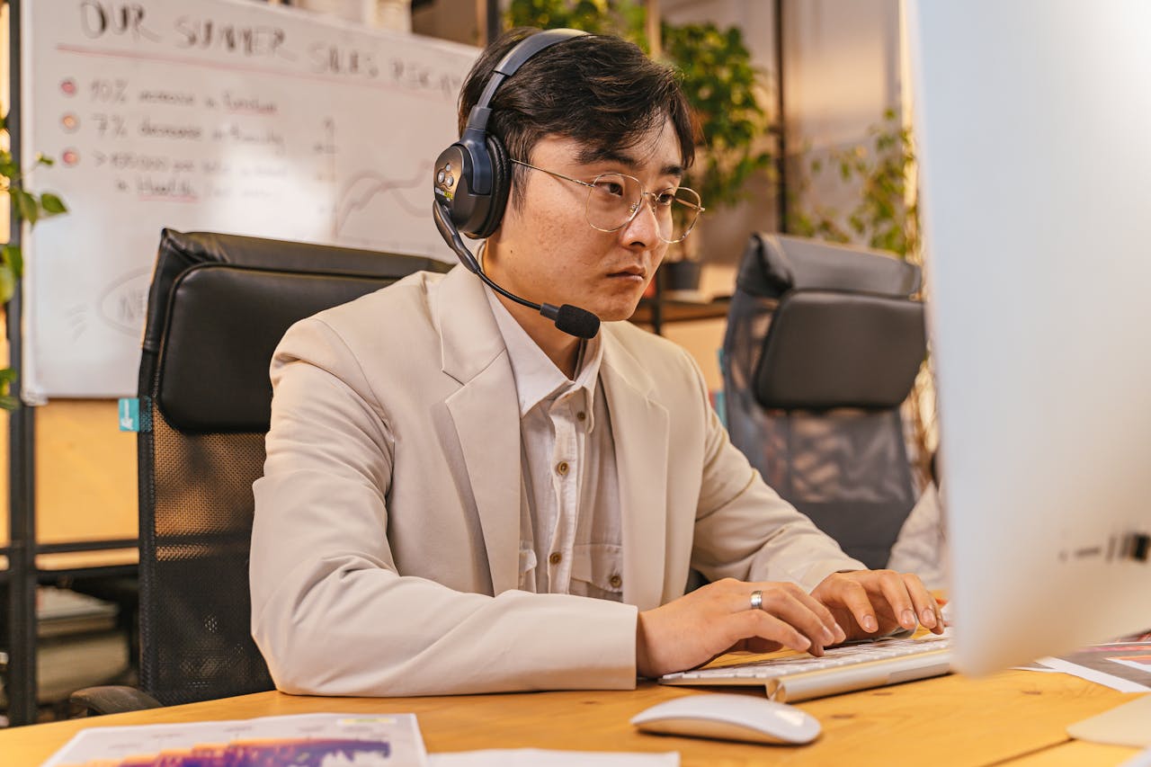 Focused man in a business suit working in office setup with headset, eyeglasses, and typing on computer.